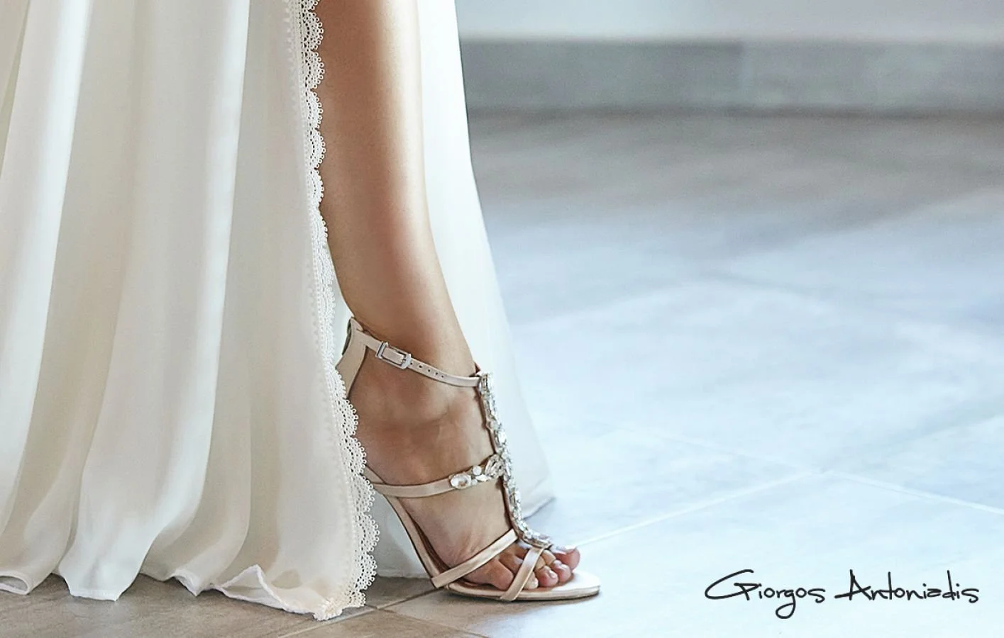 A woman's leg in a white high heel sandal, part of a white dress with lace detail, standing on a light-colored tiled floor.