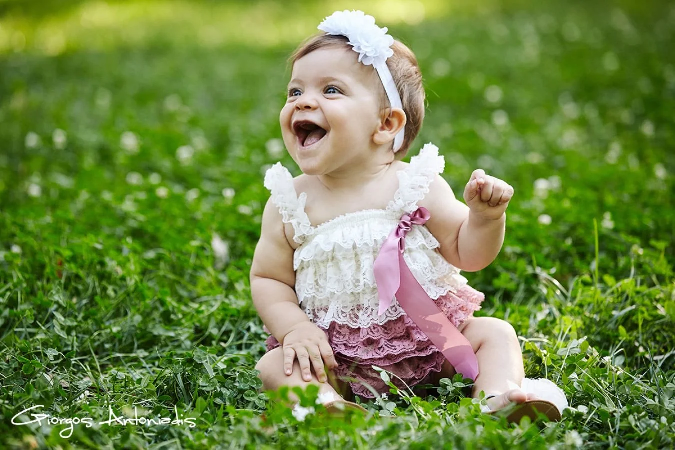 A smiling toddler girl sitting on green grass in a park, wearing a pink and white lace dress with ruffled straps, a pink ribbon, and a white flower headband.