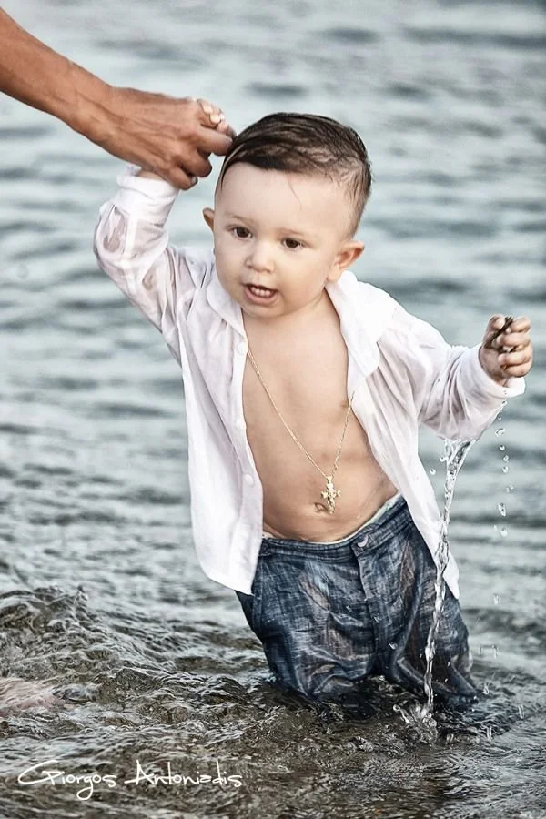 Young boy in wet clothes standing in the water at the beach, holding hands with an adult. The boy is shirtless underneath an open white shirt, wearing dark shorts, and has a gold chain around his neck.