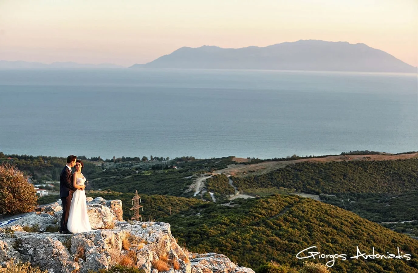 A bride and groom standing on a rocky ledge overlooking a scenic landscape with mountains and a body of water at sunset.