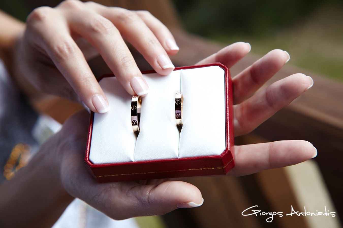 A person holding a red jewelry box with white interior, showing two rings with black and gold design. The background is blurred outdoors. The image is signed by Giorgos Antonidis.