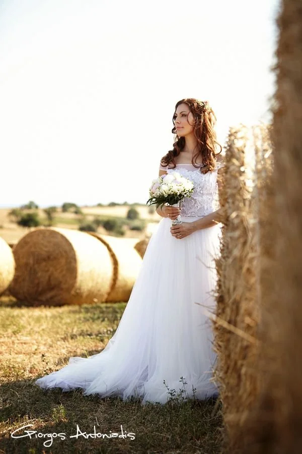 A bride in a white wedding dress holding a bouquet of white flowers, standing in a field with hay bales.