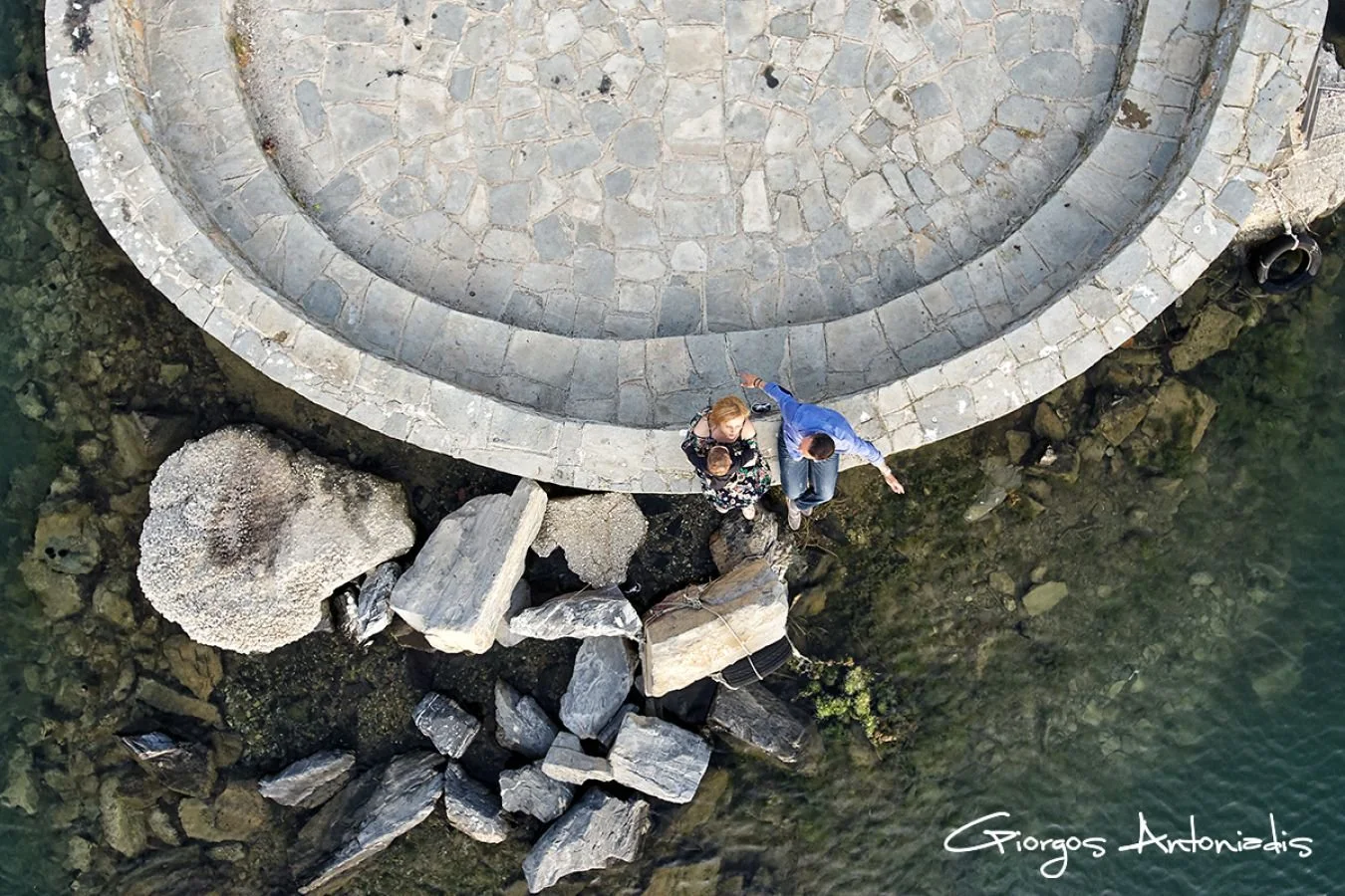 An aerial view of two people sitting on rocks by a stone pier or walkway, with water surrounding them and rocks below.
