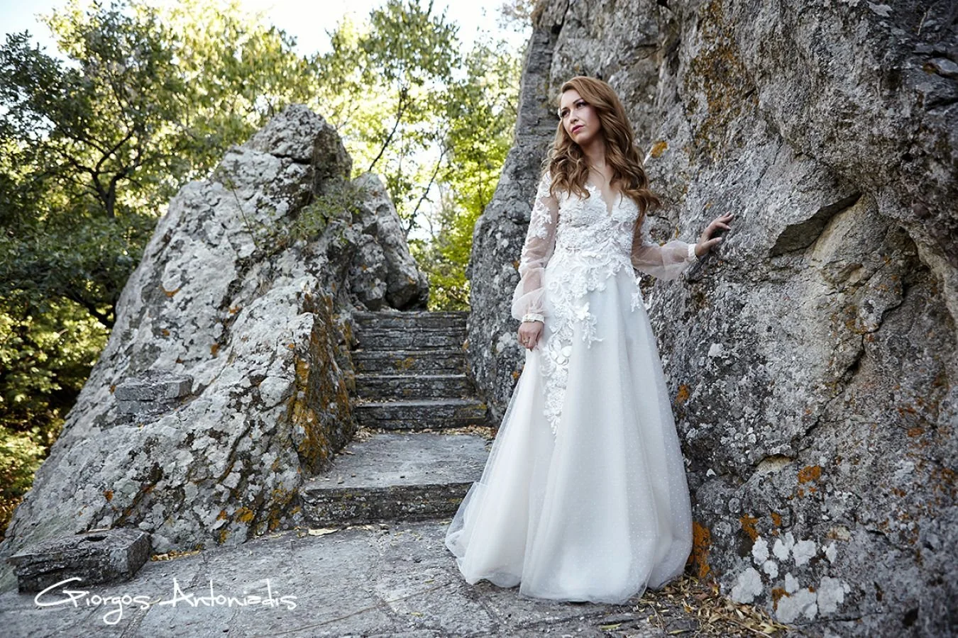 A woman in a white wedding dress standing outdoors on a rocky path, surrounded by trees and large rocks