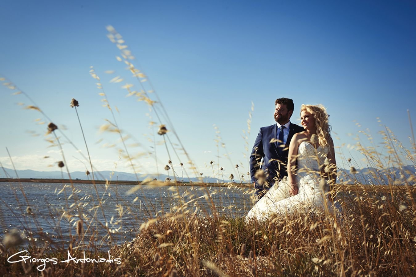 A wedding couple standing in a field with tall dry grass near a lake, with mountains in the background under a clear blue sky.