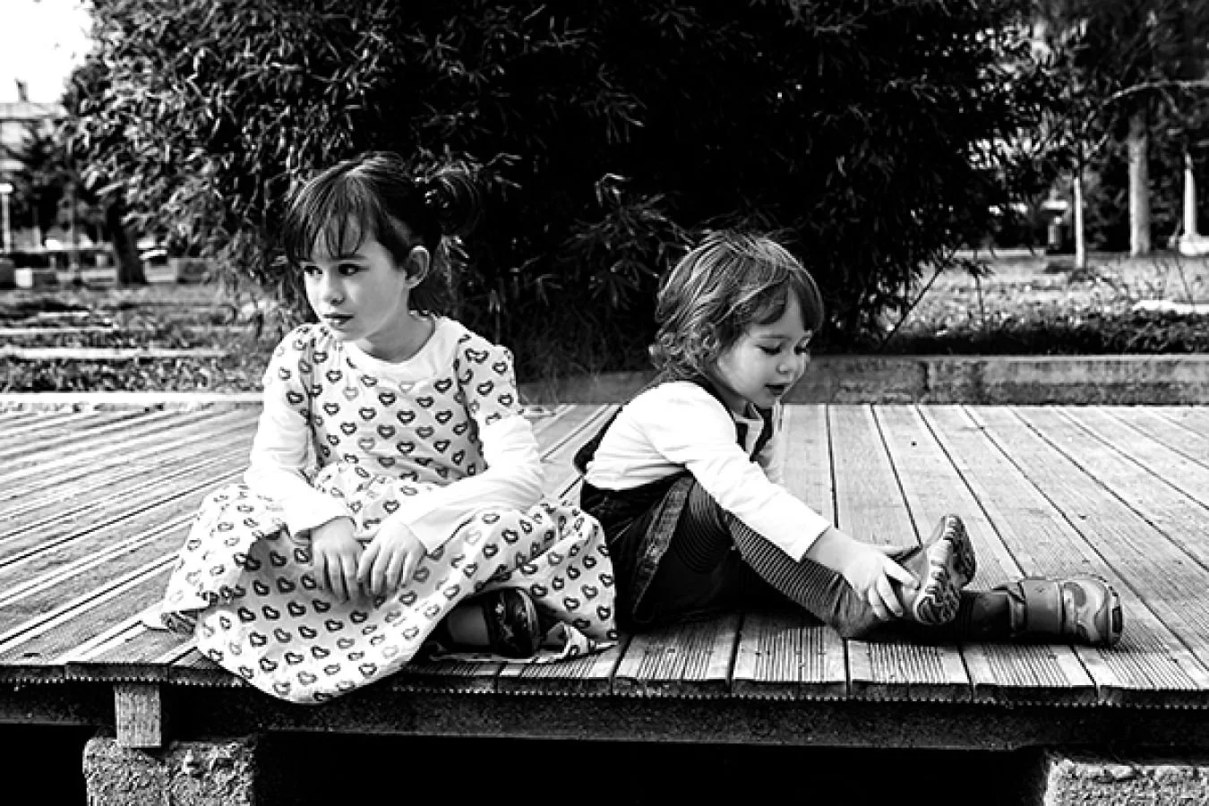 Two children sitting on a wooden deck outdoors, one girl with a patterned dress and a boy with a shirt and boots, with large bushes and trees in the background.