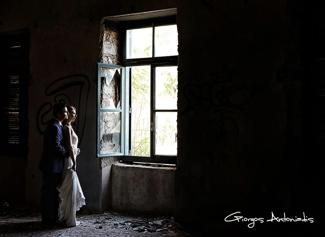 A couple dressed in wedding attire standing inside an old, abandoned building with a large open window, bright light coming through, graffiti on the wall, and a gritty floor.