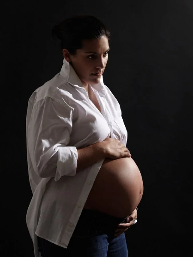 A pregnant woman with dark hair, wearing a white button-up shirt and dark jeans, stands against a dark background, gently holding her belly with one hand while looking at the camera.