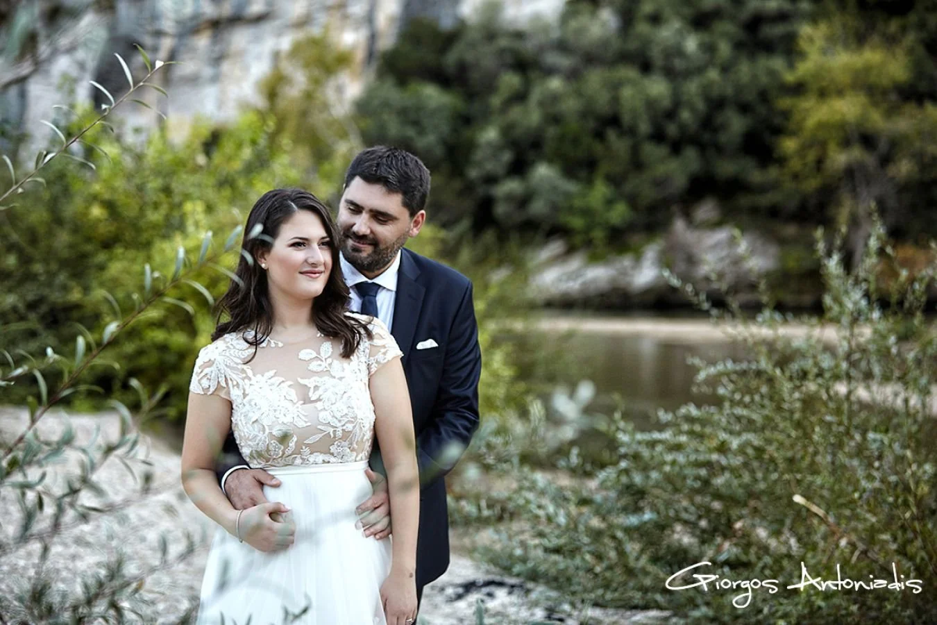A couple dressed in wedding attire standing outdoors near a body of water with greenery and trees in the background.