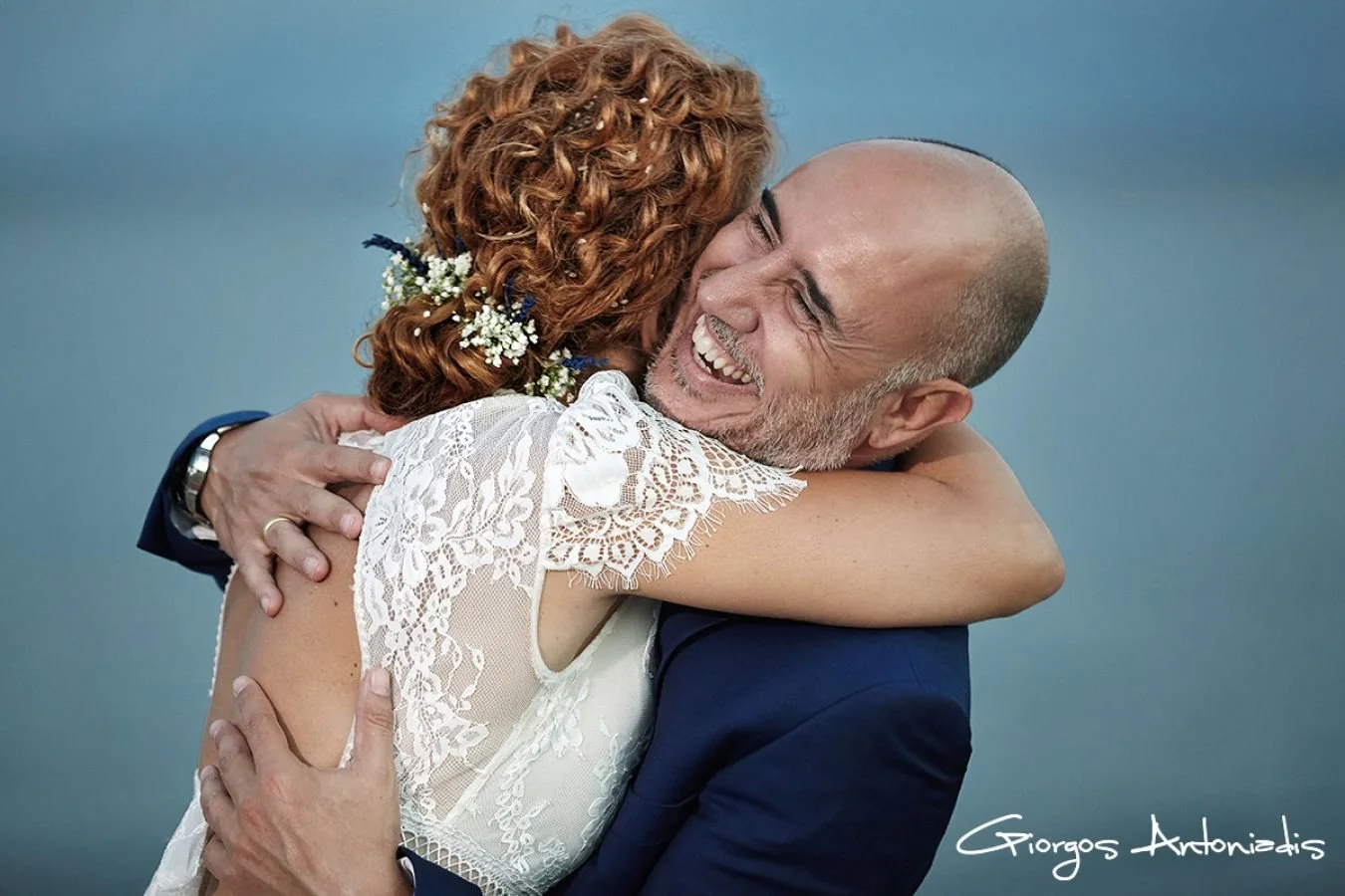 A man and a woman hugging and smiling. The woman has curly red hair with flowers, wearing a white lace dress. The man is bald with a beard, wearing a navy blue suit, embracing happily. Signature of Giorgos Antoniadis is in the bottom right corner.