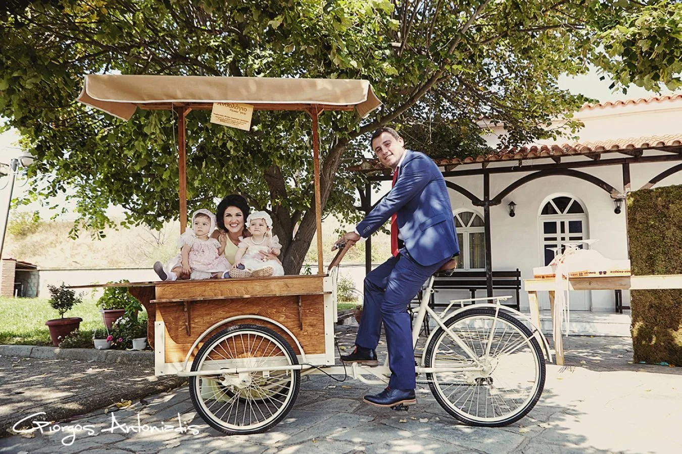 A man in a blue suit riding a white bicycle with a large wooden cart attached, carrying two young girls in pink dresses. They are outdoors, under a tree, in front of a white building with arched windows. The girl on the left is holding a finger to he