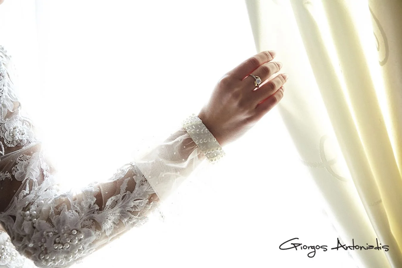 Close-up of a woman's hand with a wedding ring, touching cream-colored curtains, with a lace wedding dress sleeve visible.