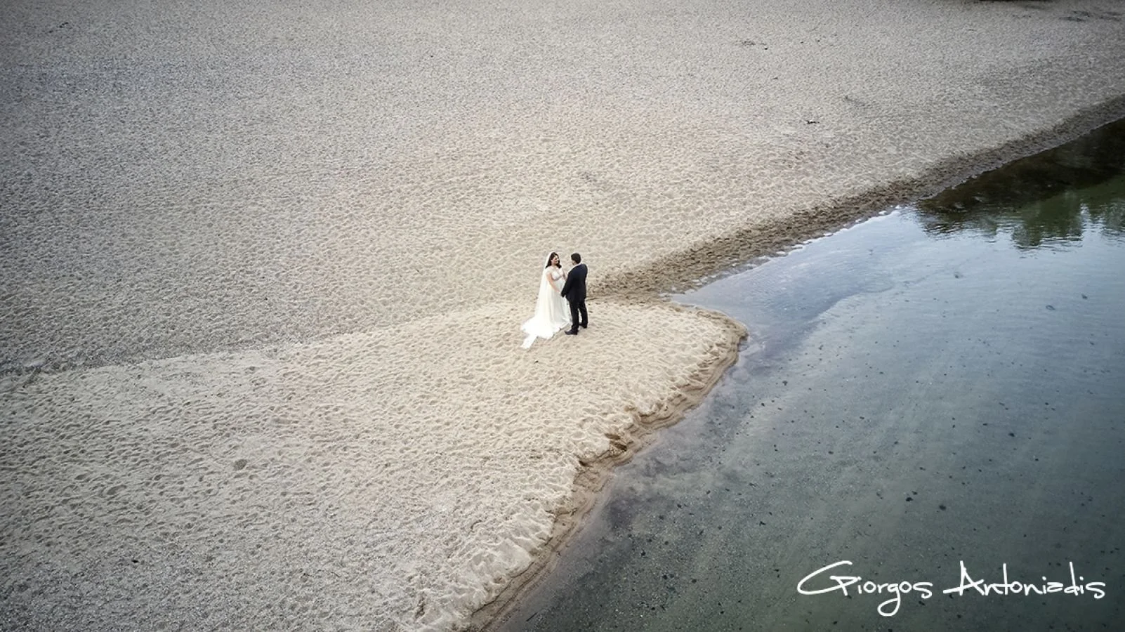 A couple dressed in wedding attire standing and holding hands on a small sand island near the water, with the bride wearing a white wedding gown and the groom a black suit, surrounded by a large sandy area and water.