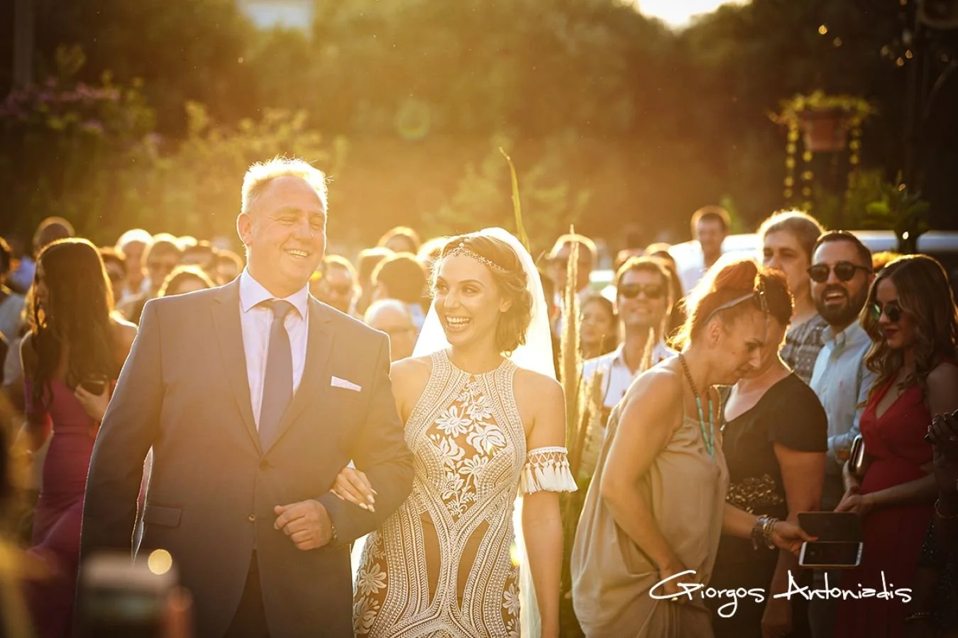 A bride and groom walking arm in arm during their outdoor wedding ceremony, surrounded by guests, with sunlight shining behind them.