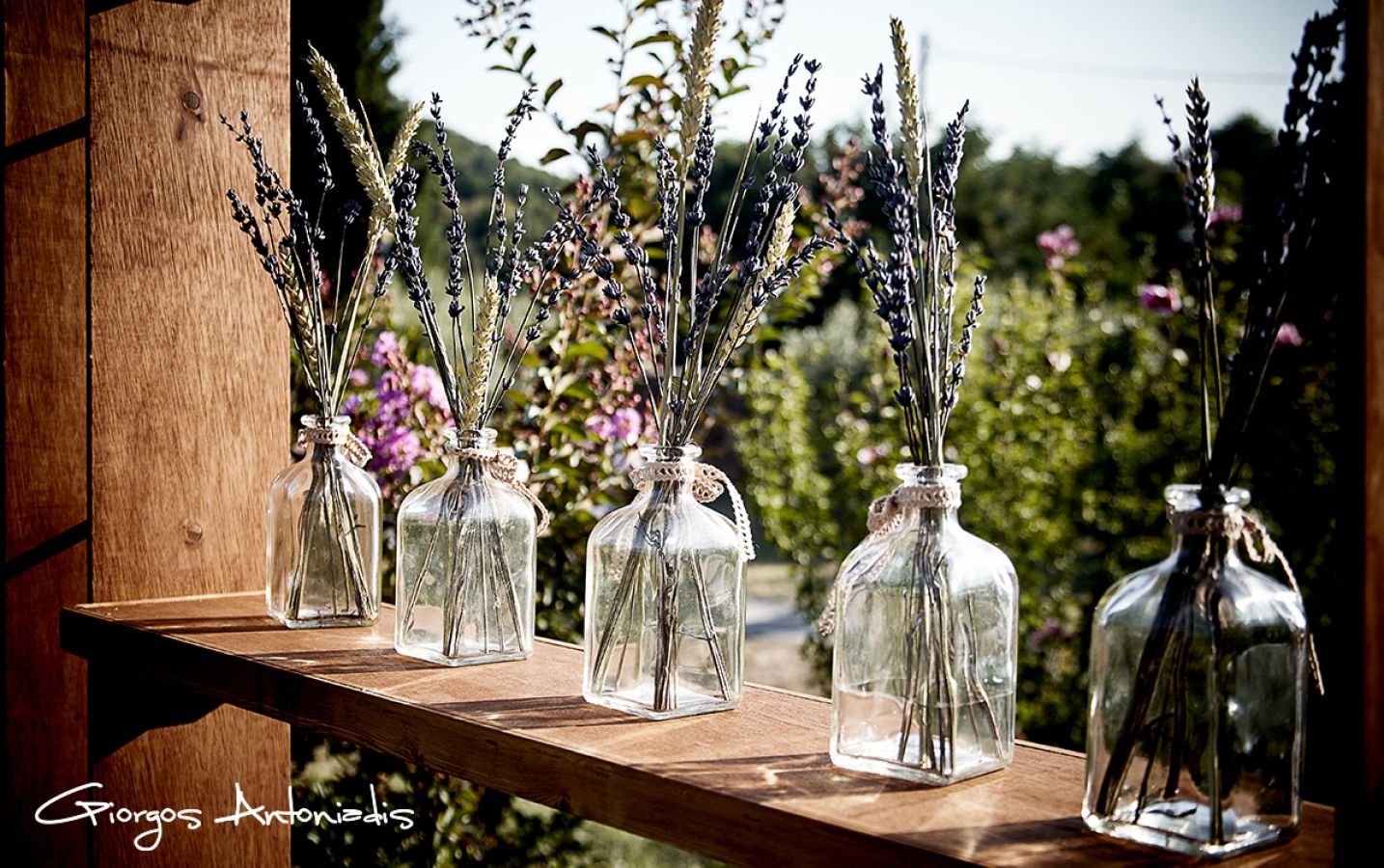 Five glass bottles with lavender and other flowers inside, arranged on a wooden ledge outdoors on a sunny day.