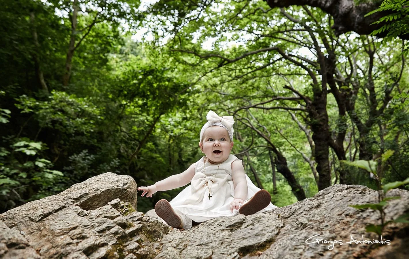 A happy baby girl in a white dress with a headband and cross necklace sitting on a large rock in a green forest.
