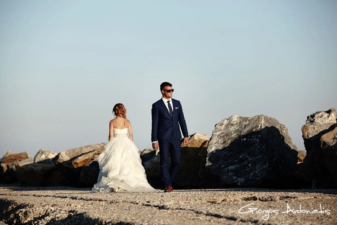 A bride in a white wedding gown with curly red hair stands with her back to the camera, while a groom in a dark suit, sunglasses, and brown shoes walks in front of her on a rocky shoreline with large rocks in the background, under a clear sky.