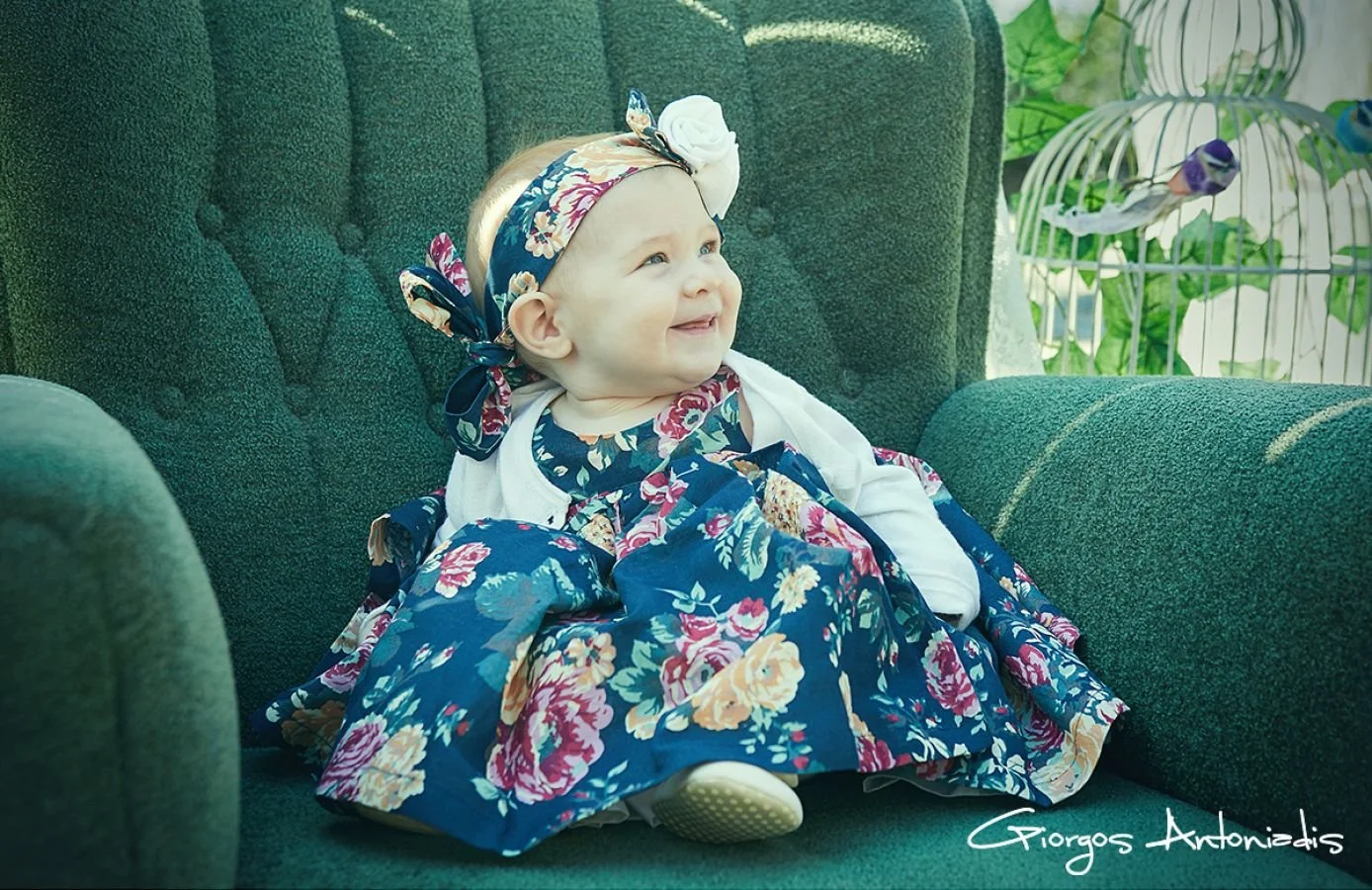A baby girl with blonde hair wearing a floral dress and matching headband, sitting on a green upholstered chair, looking happy and smiling.
