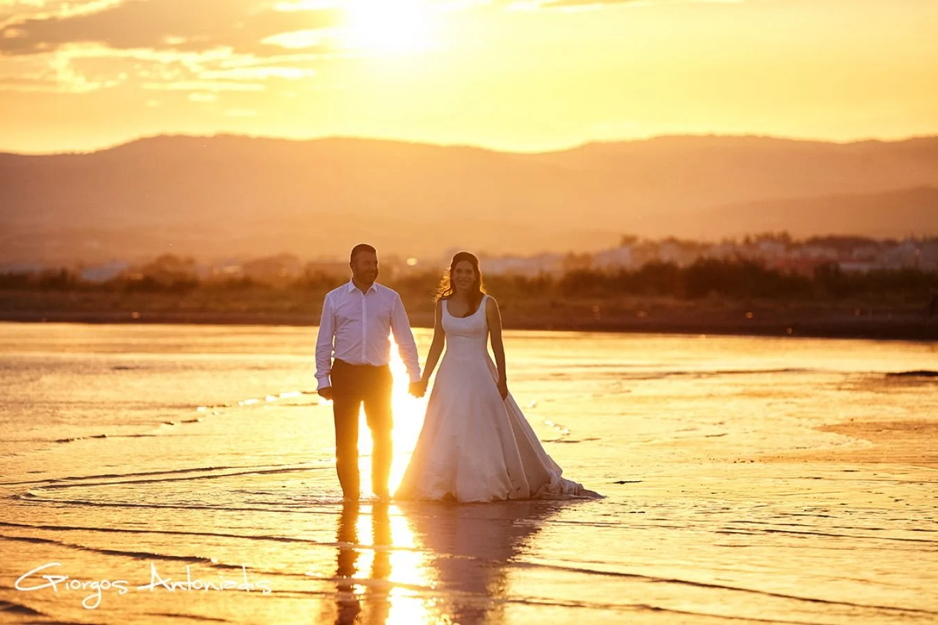 A couple in wedding attire holding hands and walking in shallow water at sunset, with mountains and sky in the background.
