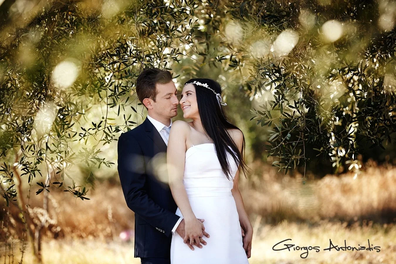 A couple dressed in wedding attire standing close together outdoors under trees, with the man in a dark suit and the woman in a white strapless dress with a floral headpiece. The woman is smiling at the man, who is looking at her with a tender expres