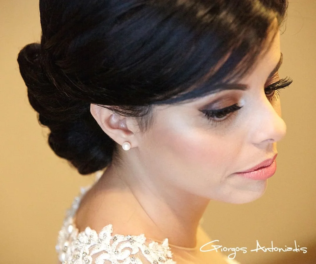 Close-up of a bride with dark hair styled in a vintage updo, wearing pearl earrings and a lace wedding dress, looking downward with a soft expression.