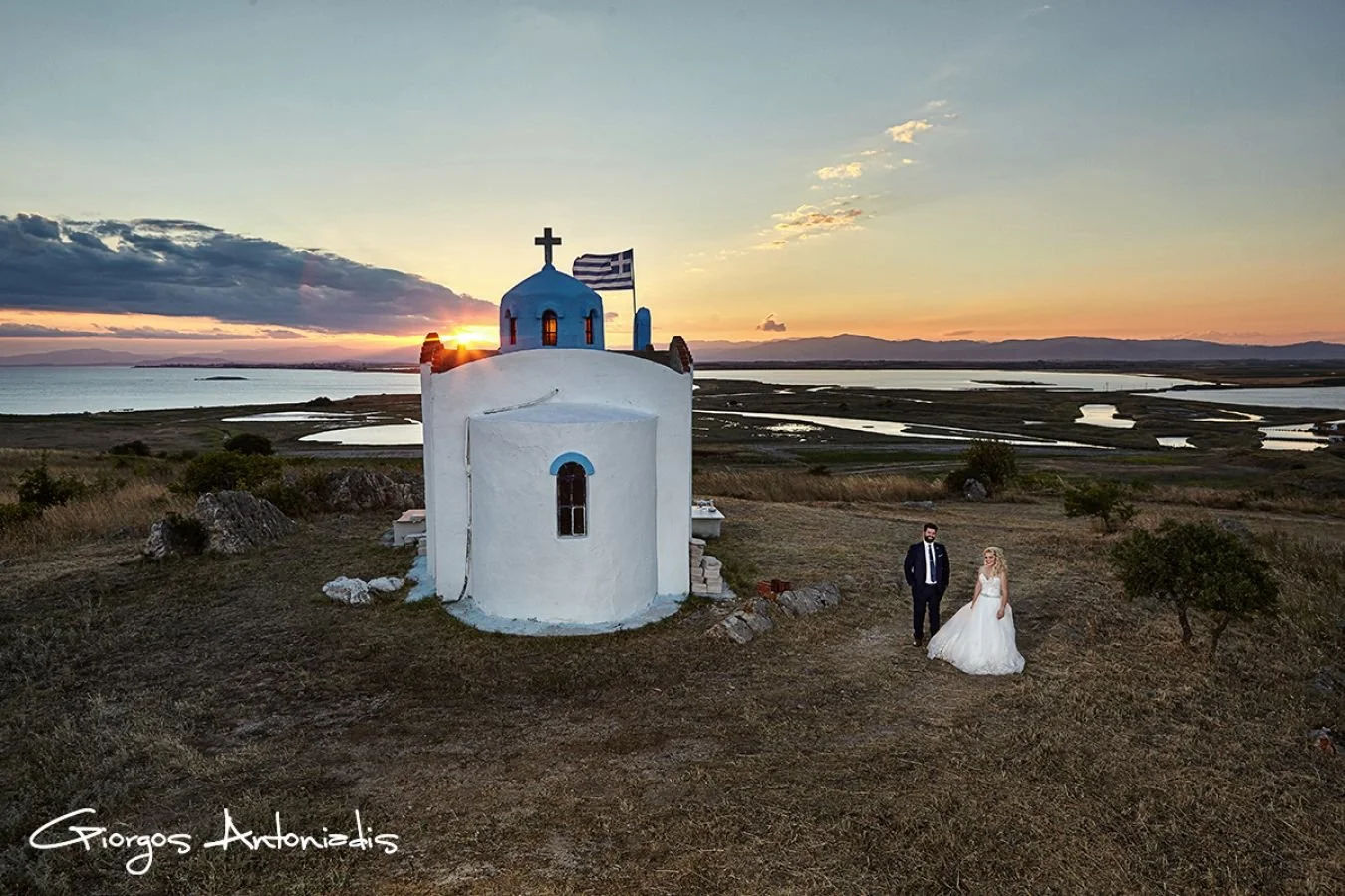 A wedding couple standing on grassy land next to a small white Greek-style chapel with a blue dome and cross, overlooking a landscape of water and hills at sunset.