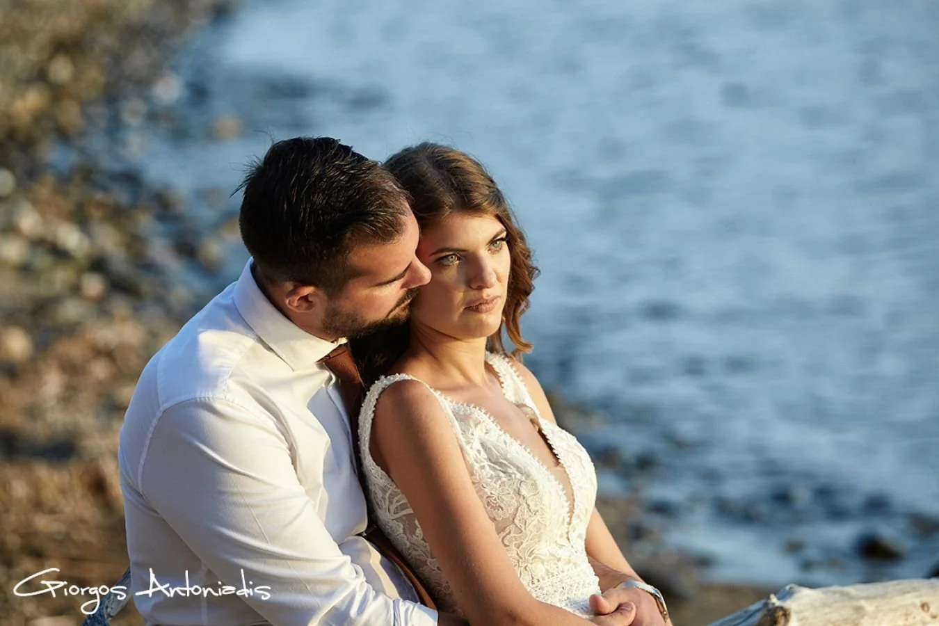 A couple sitting on a rocky beach by the water, with the man whispering to the woman who is looking away thoughtfully, during sunset.