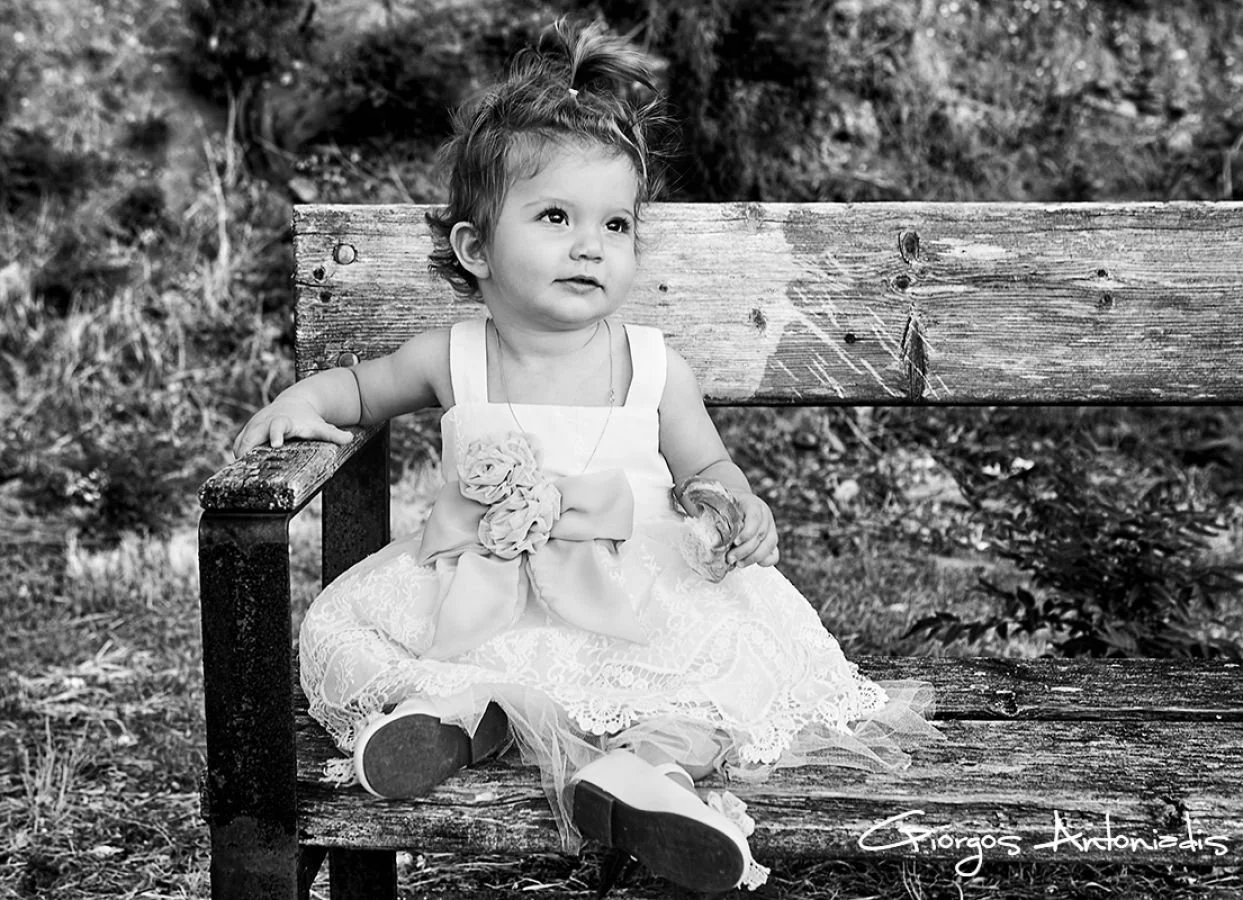 A young girl sitting on a rustic wooden bench outdoors, wearing a dress with lace details and a large bow, holding a snack, and looking thoughtful.
