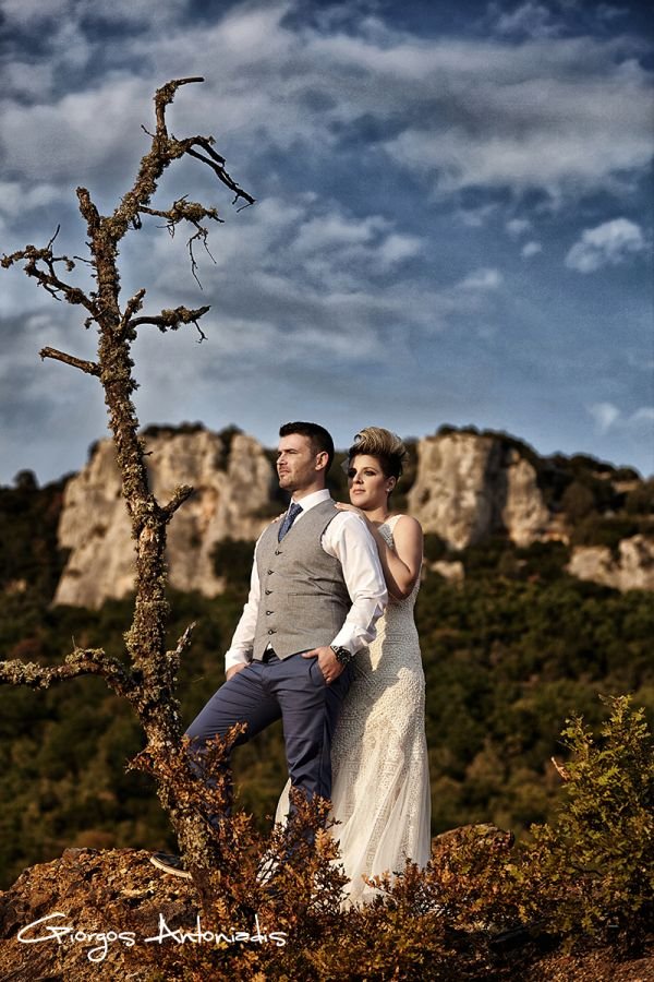 A man in a gray vest and a woman in a white lace dress standing outdoors on a rocky terrain with mountains and cloudy sky in the background.