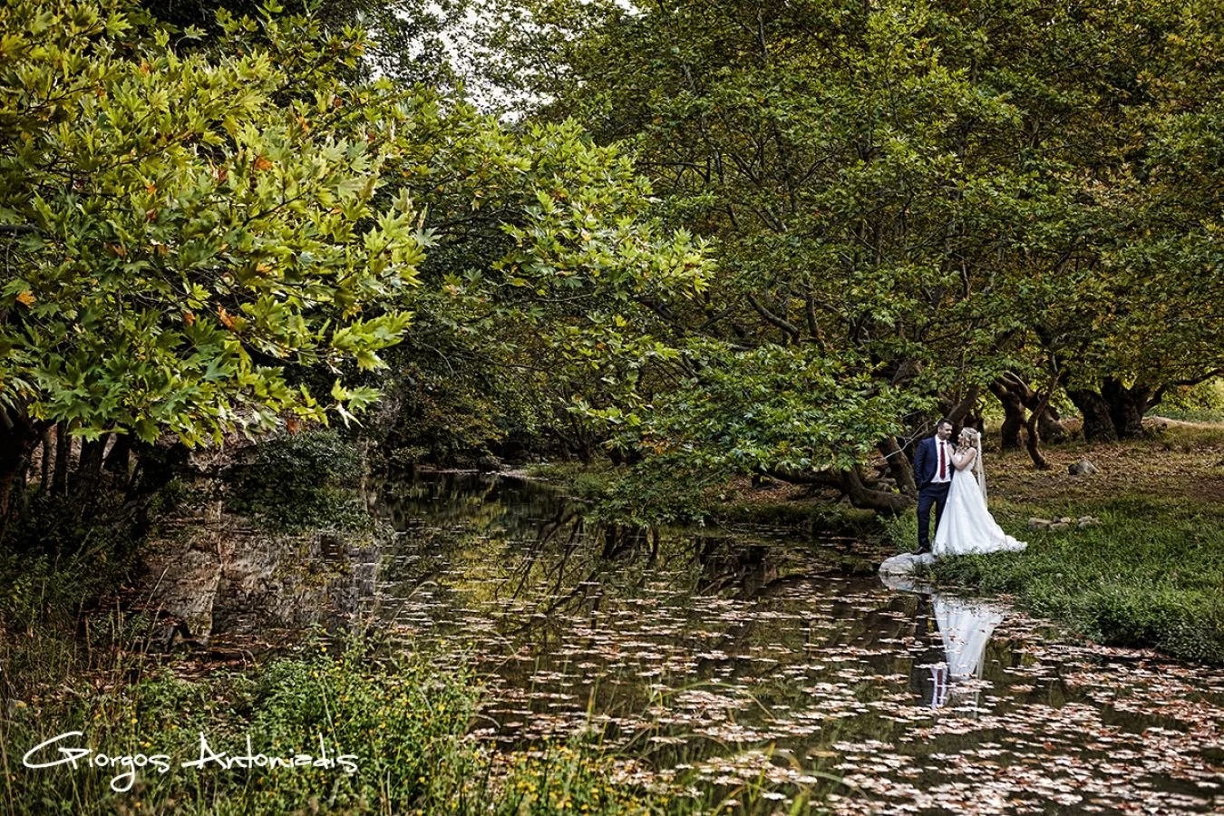 A bride and groom standing on a grassy bank beside a calm river surrounded by dense green trees, with their reflection visible in the water.