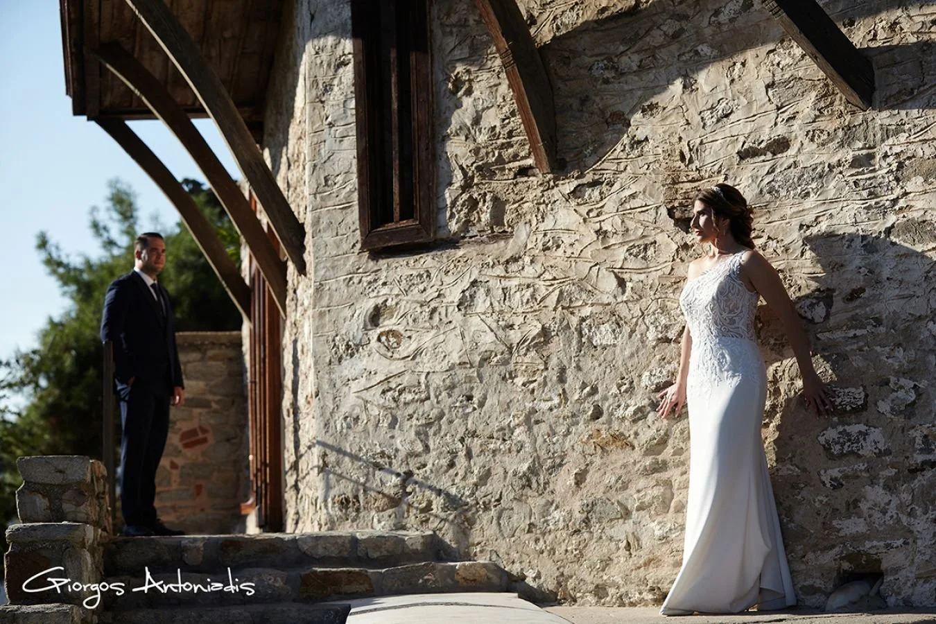 A bride in a white wedding dress standing against a stone wall, with a man in a suit standing on stairs in the background, outdoors on a sunny day.