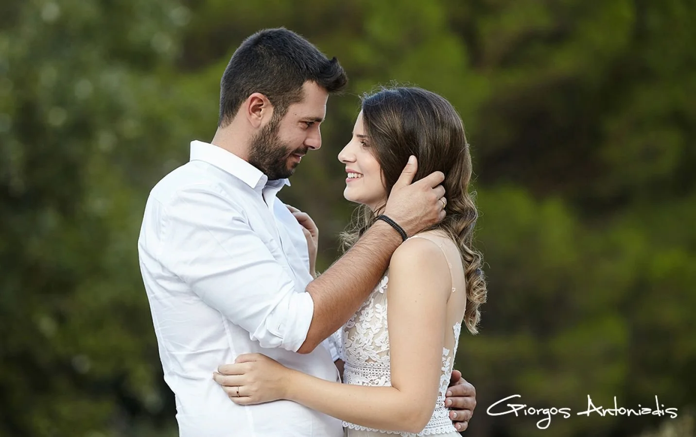 A couple gazing at each other lovingly, standing close outdoors with blurred green trees in the background.