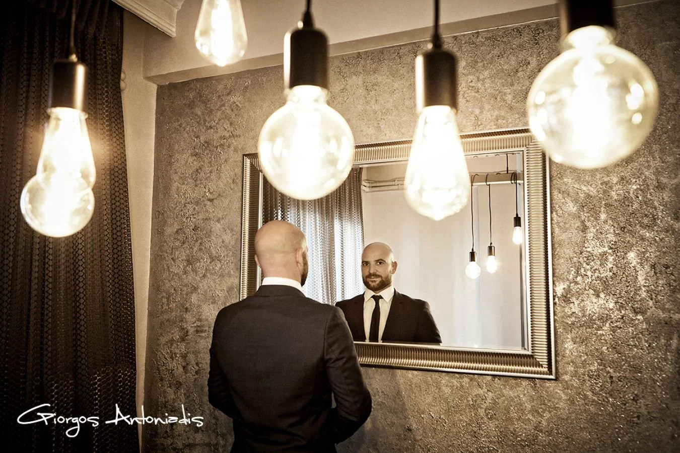 A man in a suit and tie looking at his reflection in a mirror in a warmly lit room with hanging light bulbs.