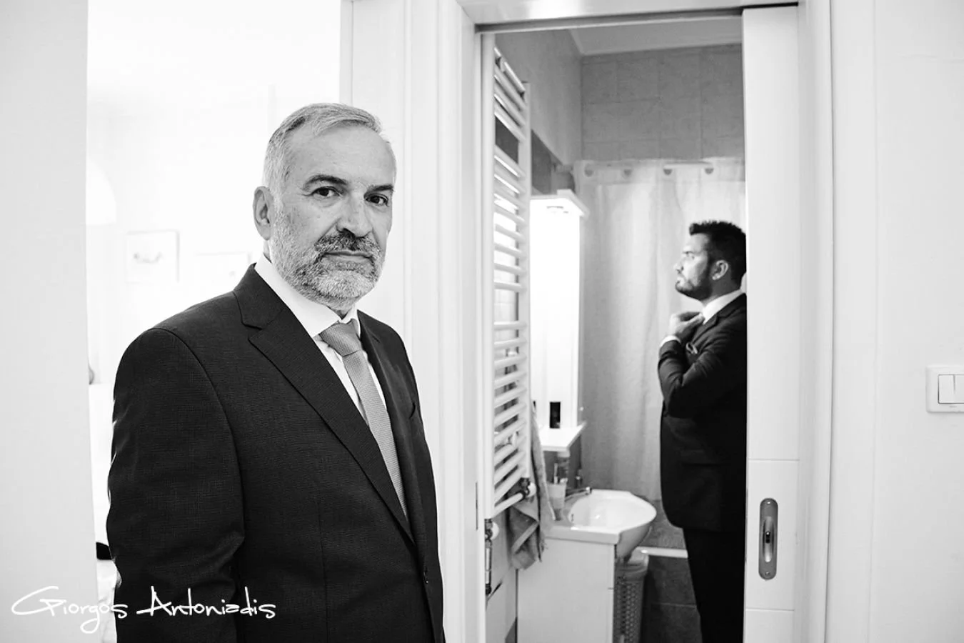 Two men in business suits, one younger and one older, in a bathroom. The older man is in the foreground, looking directly at the camera with a serious expression. The younger man is in the background, adjusting his tie in front of a mirror.