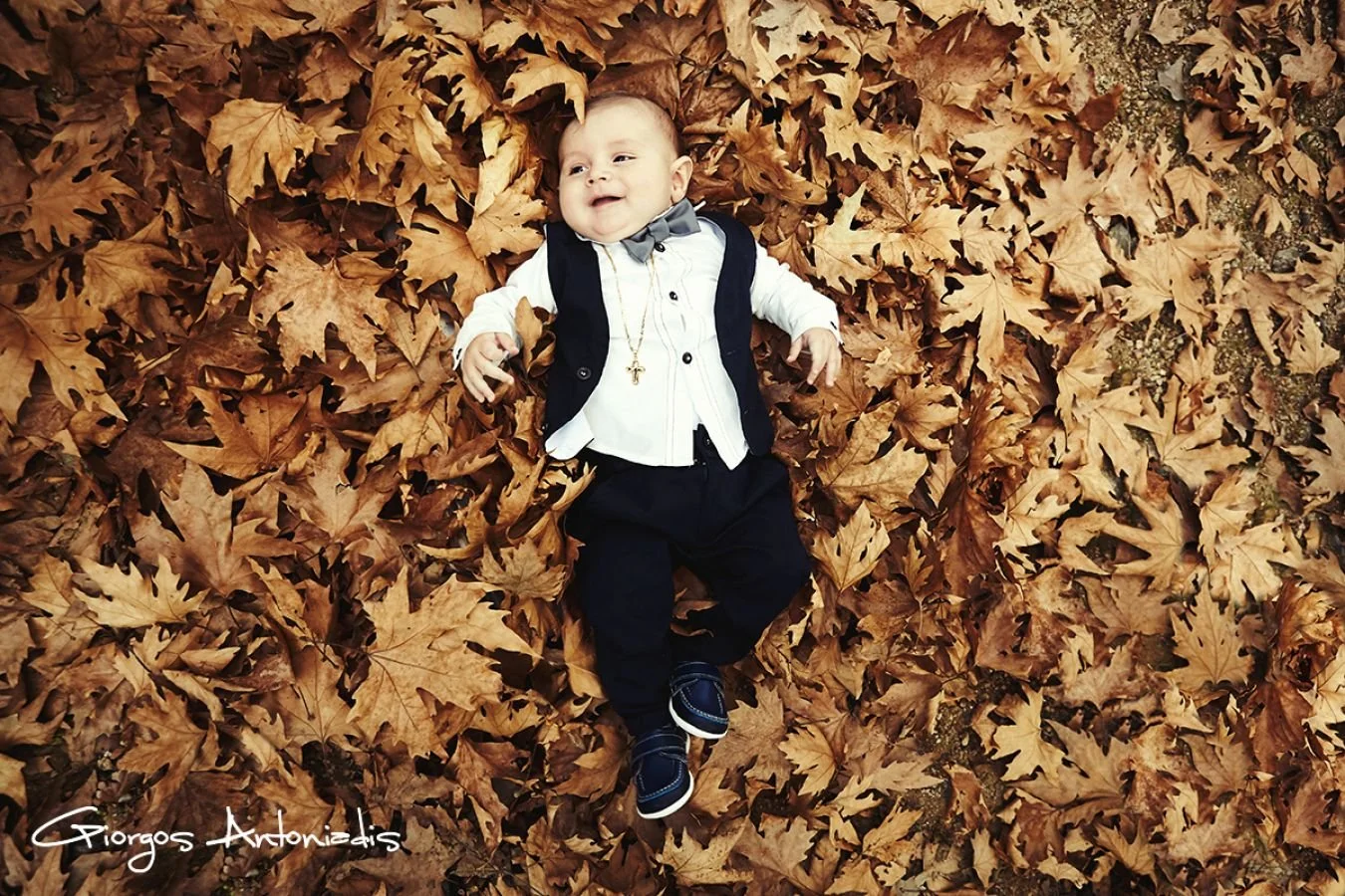 A young boy dressed in formal clothes, including a white shirt, black vest, and bow tie, lying on a blanket of autumn leaves, smiling and looking up.