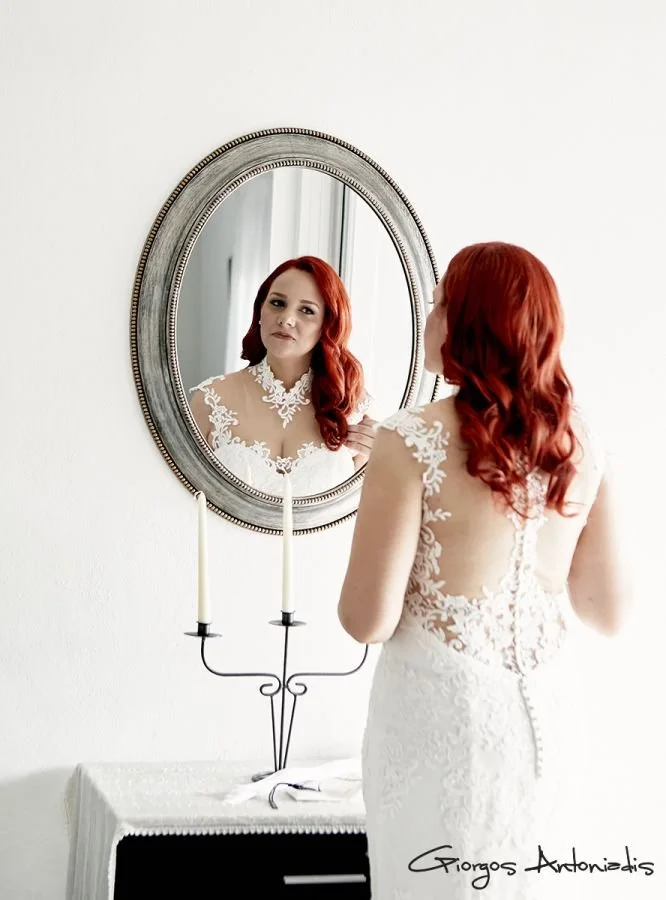 A woman with red hair in a white lace dress looks at her reflection in a large oval mirror. The mirror hangs on a white wall above a small table with a candelabra. The woman is facing away from the camera, and her reflection shows her face. The signa