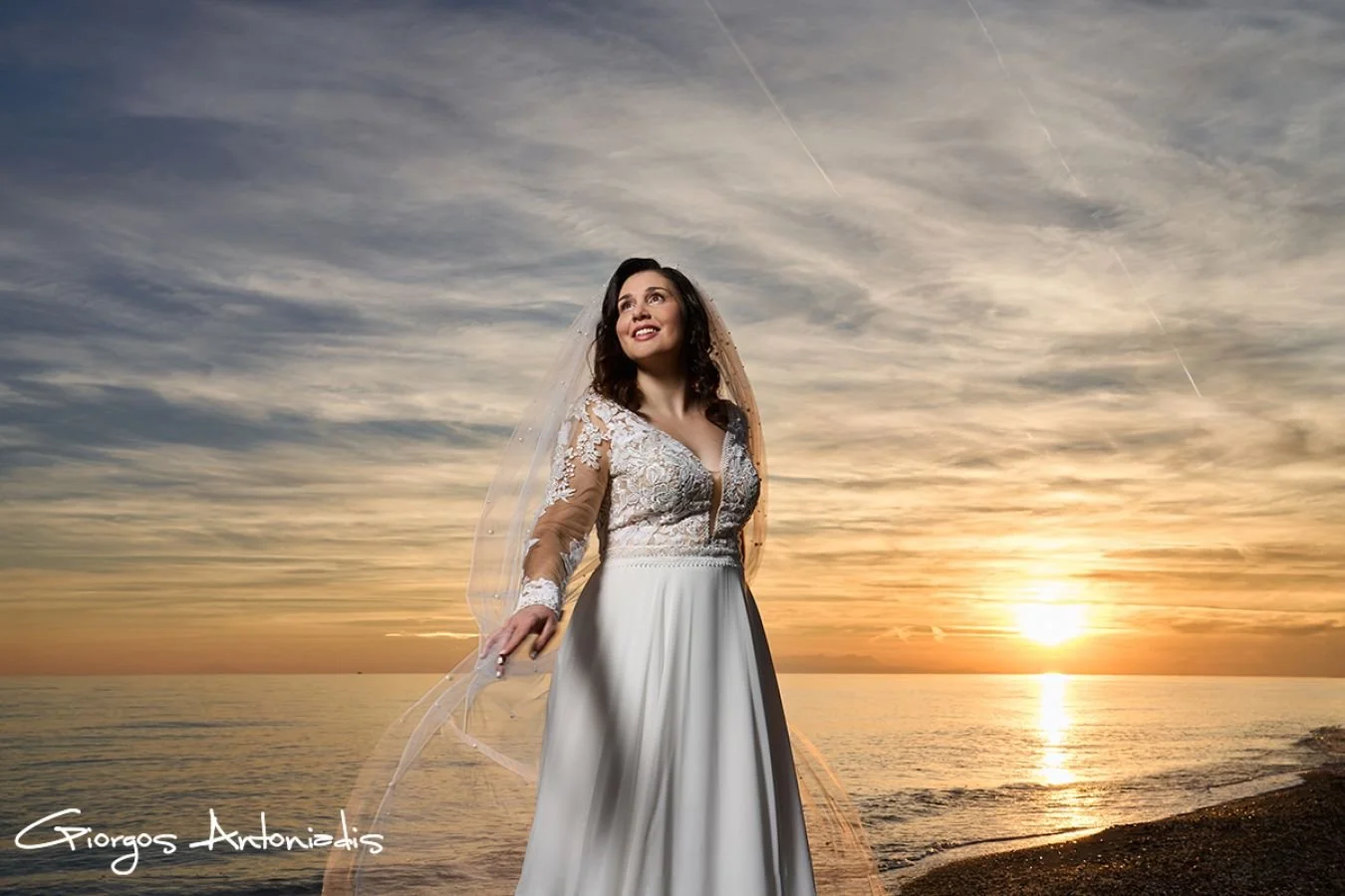 A woman in a wedding dress standing on a beach at sunset, smiling and looking up. She has dark hair and is wearing a veil. The sky is cloudy with the sun near the horizon.