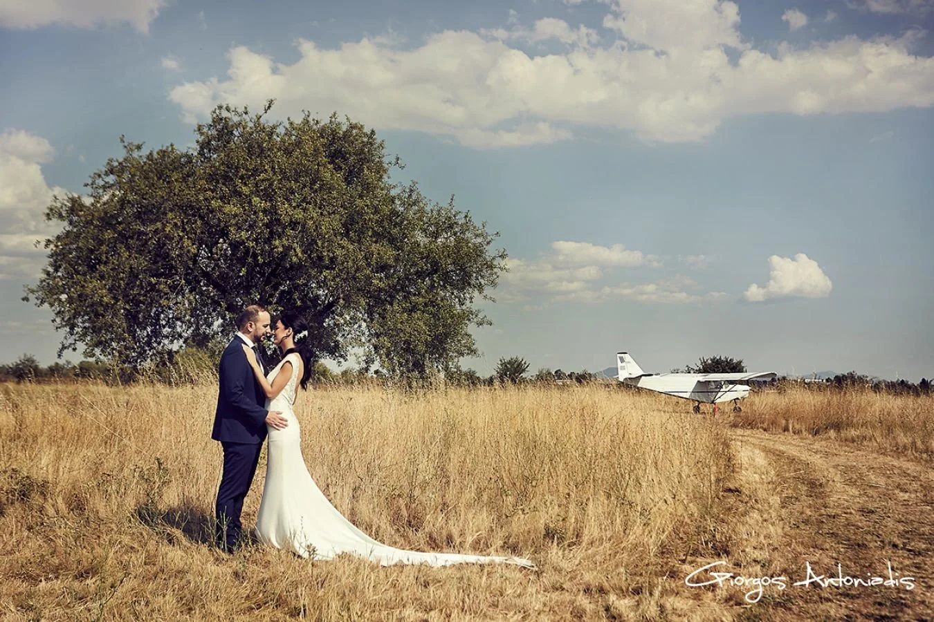 A bride and groom stand close together in a golden field, holding each other and smiling. Behind them is a large tree, and in the background are a small airplane and a blue sky with clouds. The photo is signed by Giorgos Antoniadis.