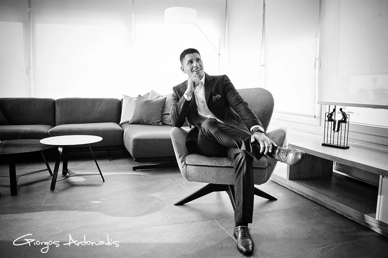 A man in a suit sitting on a modern armchair in a bright room, smiling and looking upward, with a couch and side table in the background.