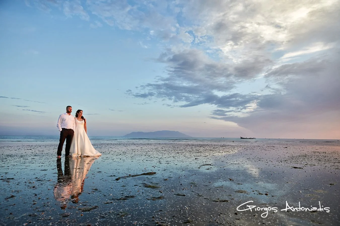A couple in wedding attire standing on a beach at sunset or sunrise, with the ocean and distant mountains in the background. The reflection of the couple is visible in the wet sand.