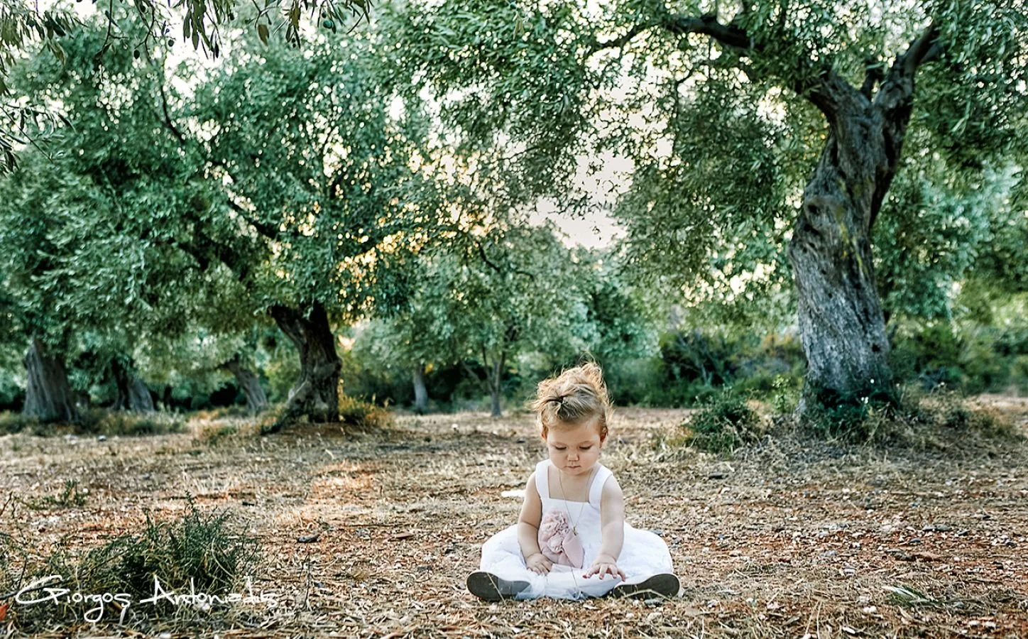 A young girl sitting on the ground in a wooded area with green trees, wearing a white dress and looking down.