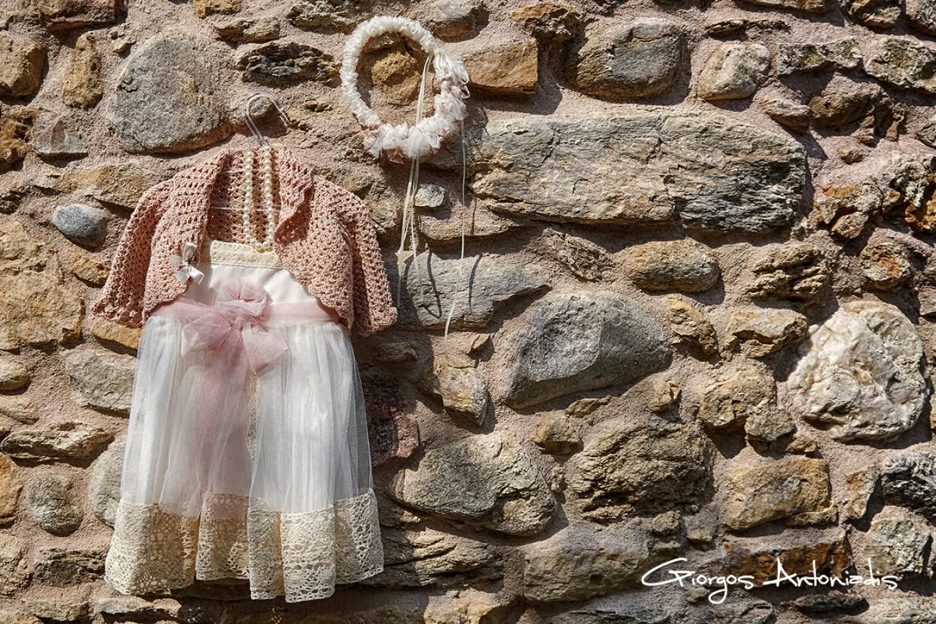 A stone wall with a doll dress hanging on a hook. The dress has a pink top, lace details, and a sheer white skirt with a pink ribbon. There is a matching headband hanging nearby.