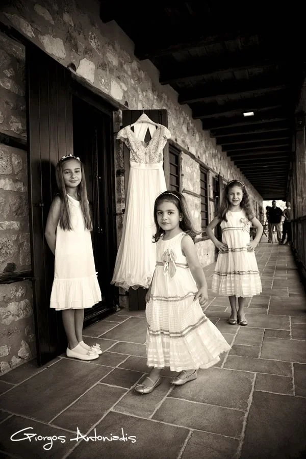 Three young girls in white dresses standing outside a rustic building, with a hanging white dress behind them.
