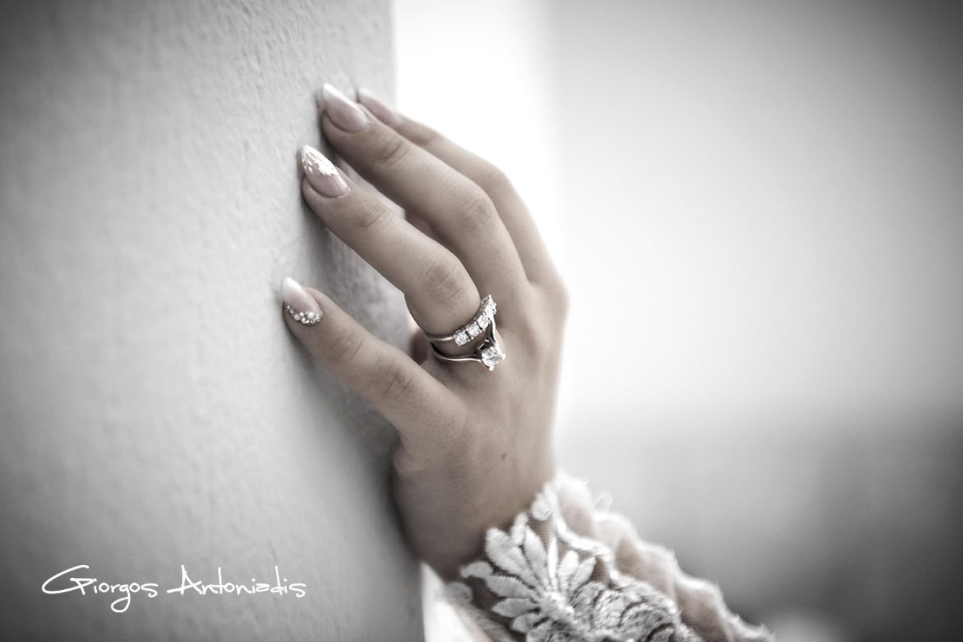 A woman's hand rests on a wall, wearing multiple rings and a lace sleeve, with natural lighting highlighting her jewelry and manicured nails.