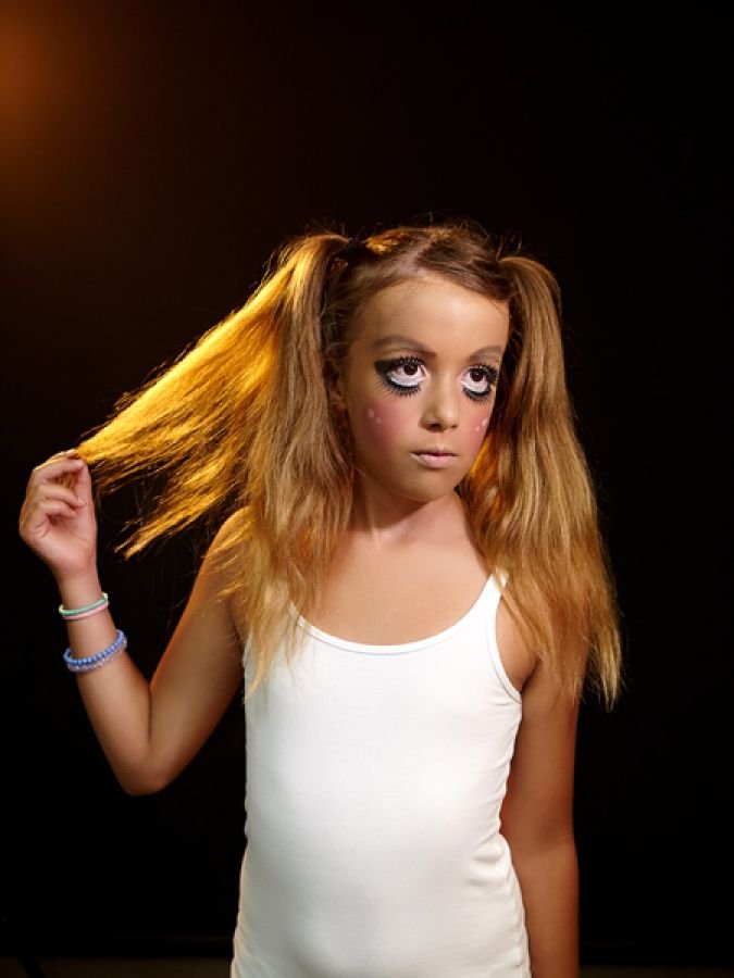 Young girl with crimped hair and dramatic makeup, wearing a white tank top, holding a strand of her hair and looking to the side against a black background.