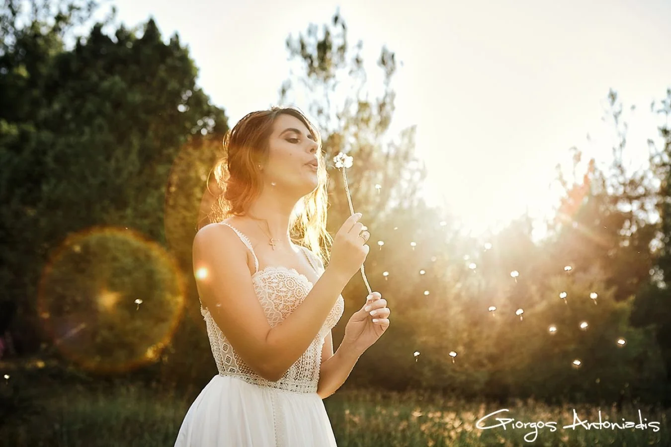A woman in a white dress blowing on a dandelion in a park during sunset, with trees in the background and sunlight causing lens flare.