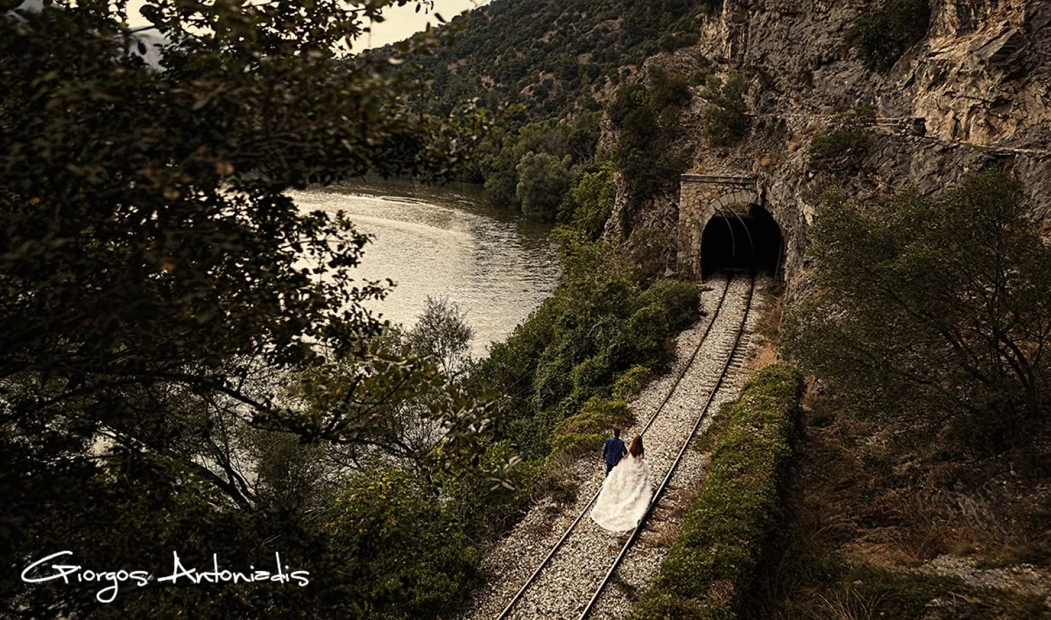 A bride and groom walking hand in hand on train tracks near a tunnel carved into a rocky hillside, overlooking a river, surrounded by trees and greenery.