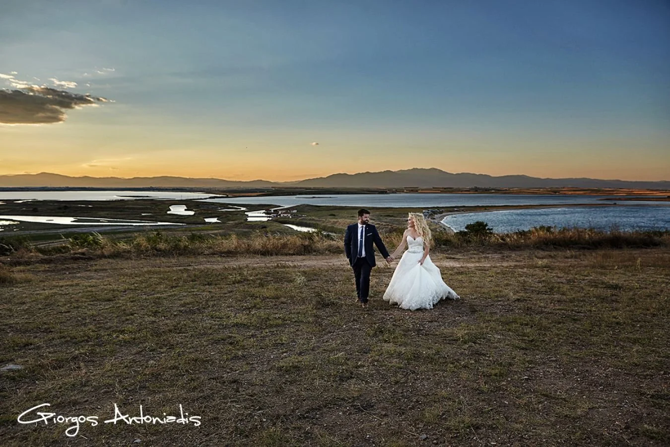 A couple holding hands on a hilltop at sunset, with a view of lakes and mountains in the background; the woman wears a white wedding dress, and the man wears a dark suit.