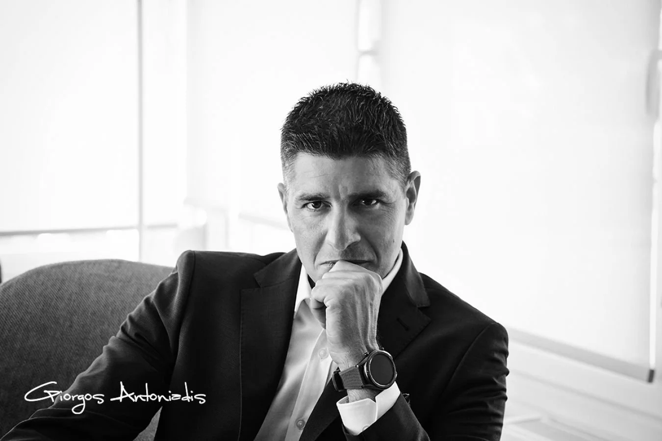 Black-and-white photo of a man in a suit sitting at a desk, with one hand resting on his chin, looking serious and contemplative.