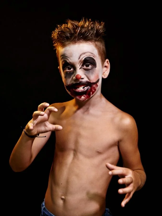 A shirtless boy with clown makeup, including dark eyes, a red nose, and a painted mouth, posing against a black background.